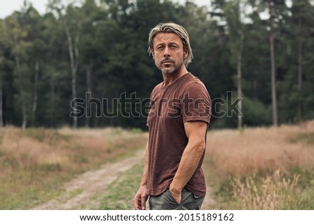 Blonde man with a stubble beard in a brown t-shirt on a sandy path in a field near a pine forest in summer. Front view.