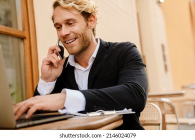 Blonde Man Talking On Cellphone While Working With Laptop In Cafe Outdoors