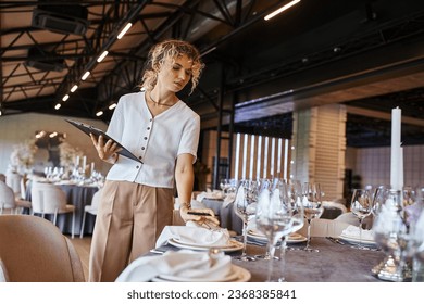 blonde manager with clipboard checking setting on festive table in event hall, banquet setup