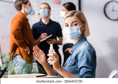 blonde manager in medical mask holding sanitizer while looking at camera near colleagues on blurred background