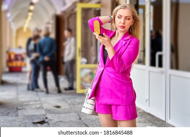 Blonde Mature Woman In Pink Suit Is Looking In The Pocket Mirror In The Trade Gallery. Mature Caucasian Female Is Making Her Make Up.