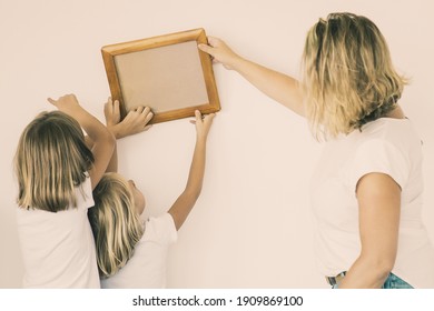 Blonde Mom And Daughters Hanging Blank Photo Frame On White Wall. Back View Of Caucasian Mother Holding And Aligning Empty Picture With Help Of Two Girls. Family, Relocation And Moving Day Concept