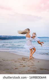 A blonde mother in a white dress got her daughter in her arms, plays with her daughter in the waves near the blue sea and laughs. - Shutterstock ID 1698830644