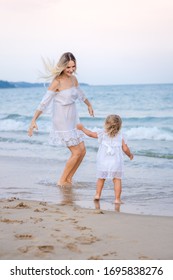 A blonde mother in a white dress plays with her daughter in the waves near the blue sea and laughs. - Shutterstock ID 1695838276
