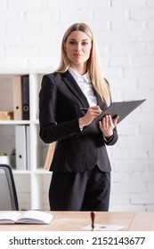 Blonde Notary With Clipboard And Pen Looking At Camera In Office