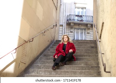 Blonde in pink fur coat sits alone on the stone steps on street in Paris