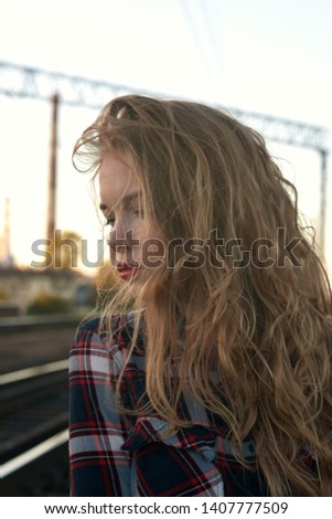 Blonde in a plaid shirt in windy weather on railway tracks. A blonde girl in a plaid shirt walks along railroad tracks in windy weather.