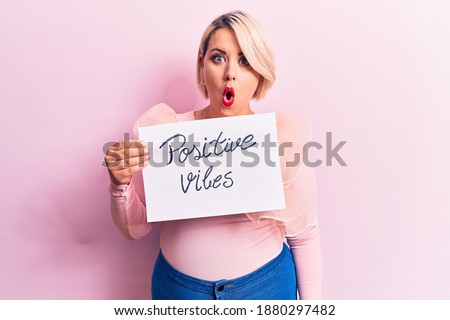 Blonde plus size woman asking for optimist attitude holding paper with positive vibes message scared and amazed with open mouth for surprise, disbelief face