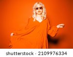 The blonde poses on a yellow background in stylish orange glasses and a dress. Girl model in the studio, bright photo of fashion and style, gold jewelry 80s and 90s. Place for text.