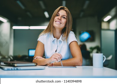 Blonde positive young woman dressed in casual wear smiling while sitting at table with modern touch pad device with stylus in hand.Cheerful student preparing for seminar using table and electronic pen