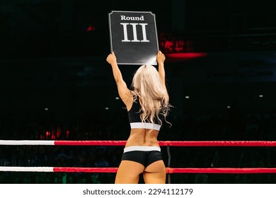 blonde ring girl demonstrate sign with number third round during fight MMA on dark background