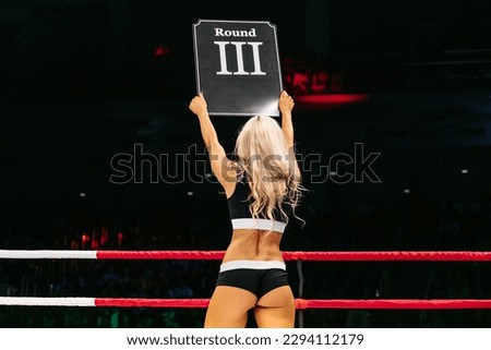 blonde ring girl demonstrate sign with number third round during fight MMA on dark background