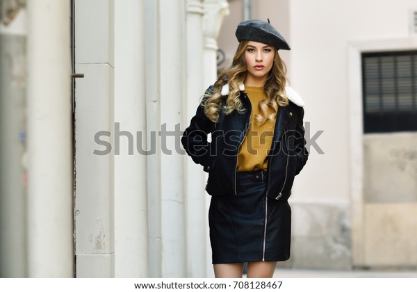 Blonde russian woman in urban background. Beautiful\
young girl wearing beret, black leather jacket and mini skirt\
standing in the street. Pretty female with long wavy hair hairstyle\
and blue eyes.