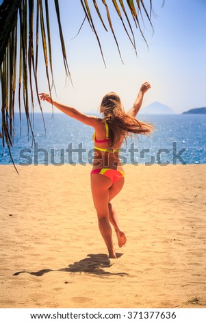 blonde slim female gymnast in bikini backside runs barefoot to sea hands up behind palm branch wind shakes long hair