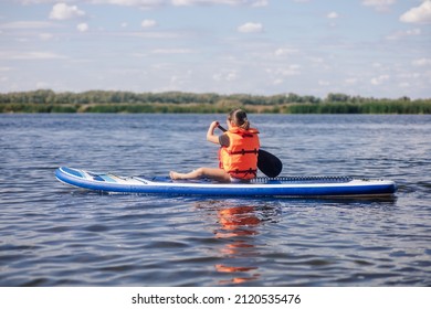 Blonde small girl sup boarding on lake in daytime rowing with oar with reeds and trees in background wearing vest life jacket. Active holidays. Inculcation of love for sports from childhood. - Shutterstock ID 2120535476