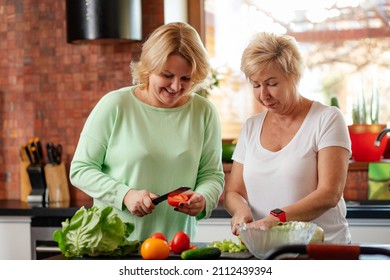 Blonde Smiling Senior Woman And Daughter Help Cook Dinner Together In Light Kitchen. Family Grandmother Teach Housekeeping Prepare Healthy Food Vegetable Lunch Two Persons Lifestyle.