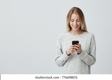 Blonde smiling young woman demonstrating white teeth using cell phone, messaging, being happy to text with her boyfriend, looking at screen of smartphone. Modern technologies and communication - Powered by Shutterstock