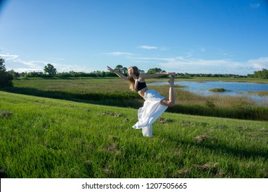 Blonde Spiritual Woman Practicing Yoga And Doing The King Dance Standing Pulling Bow Or Natarajasana / Dandayamana Dhanurasana Pose In A Park - Model Wearing Long White Flared Hippie Pants