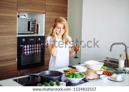 Blonde student girl standing at the table with food ingredients placed in line, going to make dinner at home kitchen and calling to her female friends, inviting to taste her healthy dish.