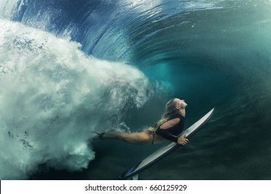 A blonde surfer girl underwater doing duck dive holding surfing board left behind air bubbles in blue water background under big ocean wave