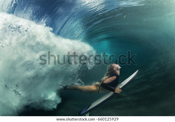 A blonde surfer girl underwater doing duck dive\
holding surfing board left behind air bubbles in blue water\
background under big ocean\
wave
