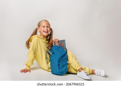 A Blonde Tanned 7 8 Year Old School Girl In Yellow Sweatshirt Jump Suit With A Blue Bag Rucksack In Hands Is Sitting And Smiling Ready To Start School Day. Isolated On Grey Background, Free Copy Space