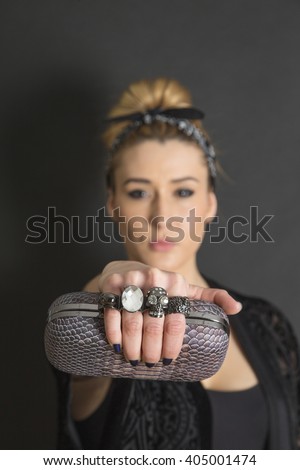 Blonde tattooed fashion girl  with bun and handkerchief holding a handbag with many rings on black background