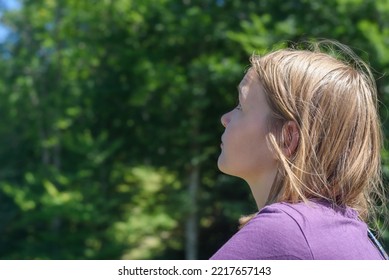 Blonde Teen Girl Explores Destinations At Back Sunlight. Teenager Tourist Looks Around Standing Against Blurry Green Trees At Resort