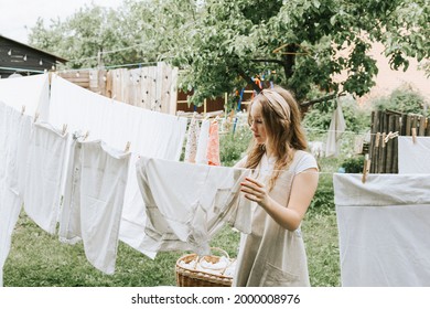 blonde teen girl in linen apron is doing homework washing clothes in basin and hanging them on ropes on the street in courtyard of village cottage house, concept of summer and freshness, laundry day