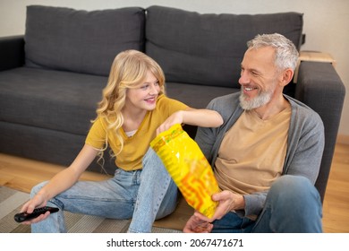 A Blonde Teen Sitting On The Floor With Her Dad And Eating Chips