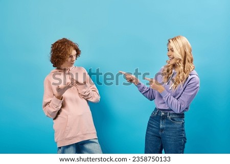 blonde teenage girl laughing and pointing with fingers at discouraged redhead boyfriend on blue