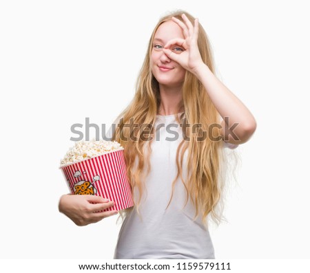 Blonde teenager woman eating pop corn with happy face smiling doing ok sign with hand on eye looking through fingers