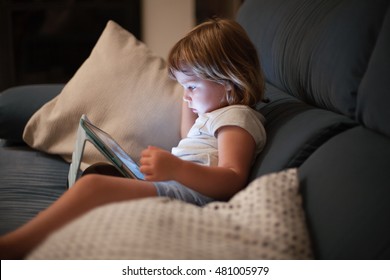 blonde three years old baby shirt and shorts, sitting comfortably in sofa inside home at night reading and watching digital tablet, face illuminated by the light of the screen - Powered by Shutterstock