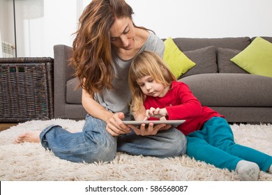 Blonde Three Years Old Child And Mother, With Red, Green And Grey Clothes, Touching And Watching Digital Tablet To Surf Internet, Sitting On Carpet Indoor Home