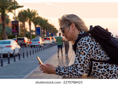 A blonde tourist girl in the Dubai resort area wanders around, taking selfies against the magnificent backdrop of modern architecture, lively harbor, opulent boats, towering skyscrapers, 