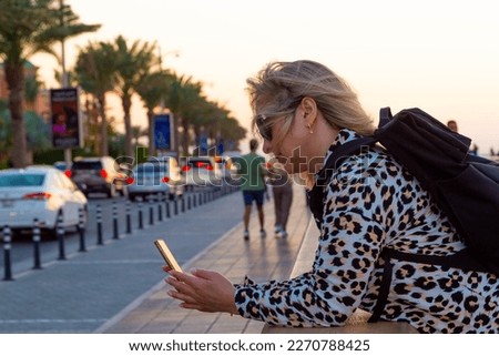 A blonde tourist girl in the Dubai resort area wanders around, taking selfies against the magnificent backdrop of modern architecture, lively harbor, opulent boats, towering skyscrapers, 
