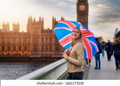 A blonde tourist woman with a british flag umbrella stands in front of the Big Ben Tower in London, Westminster, during a sightseeing trip through the city, United Kingdom