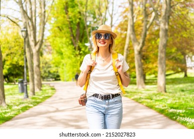 A Blonde Tourist Woman Smiling With A Hat And Sunglasses Walking In The Spring In A Park In The City, With The Camera, Enjoying The Sun