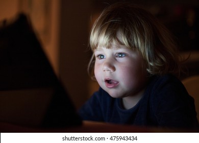 Blonde Two Years Old Baby Blue Sweater, Sitting Inside Home At Night Reading And Watching Digital Tablet, Face Illuminated By The Light Of The Screen Astonished