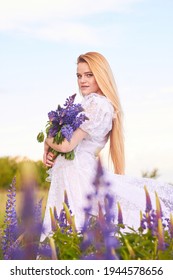 blonde in white vintage dress and bouquet lupins among purple field 
