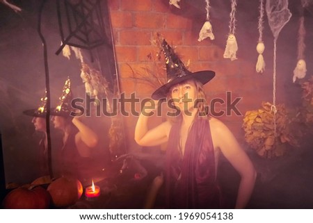 Blonde witch in red dress and black hat with candle and mirror in Halloween decoration indoors