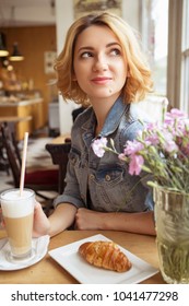 Blonde (with dark roots) caucasian woman in casual summer outfit at the cafe. Grey dress and jeans jacket. Woman got natural day makeup and curly hairstyle. She drinks coffee latte and eats croissant