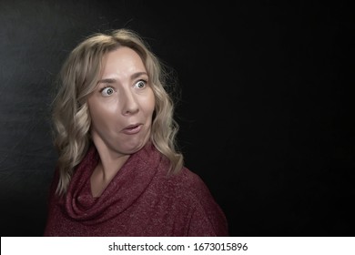A blonde woman 35-40 years old with an emotion of strong surprise on her face on a dark background, a place for text.