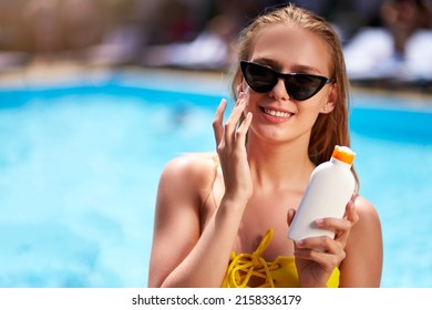 Blonde woman applies sunscreen solar cream on cheeck and face near swimming pool. Smiling pretty girl puts suntan cream from plastic bottle on skin in spa. Female in bikini with suntan lotion.