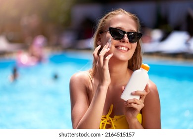 Blonde woman applies sunscreen solar cream on cheeck and face near swimming pool. Smiling pretty girl puts suntan cream from plastic bottle on skin in spa. Female in bikini with suntan lotion. - Powered by Shutterstock