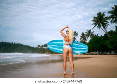 Blonde woman in bikini holding surfboard on tropical beach, preparing for surfing session. Female surfer enjoying summer vacation by the sea, getting ready to catch waves under sunny sky. - Powered by Shutterstock