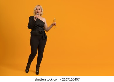blonde woman in black clothes with bare shoulder holding a microphone and a glass of champagne on a colored background with copy space