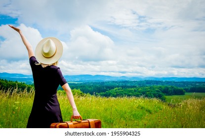 Blonde woman in black dress and suitcase at meadow with mountains on background - Shutterstock ID 1955904325