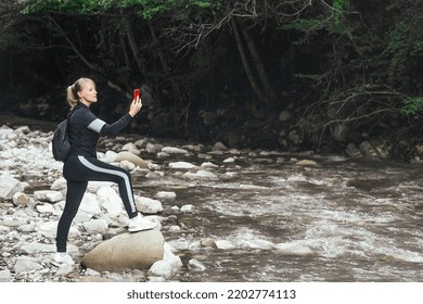 a blonde woman blogger in sports comfortable clothes and with a backpack on her shoulders takes pictures of nature in the mountains and a mountain river with a course on her phone - Shutterstock ID 2202774113