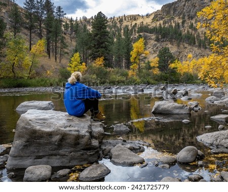 Blonde woman in blue sweatshirt sitting on boulder in the river.  Autumn color and mountain in the background with hint of blue sky. Whitecaps on river flowing swiftly with trees in background.